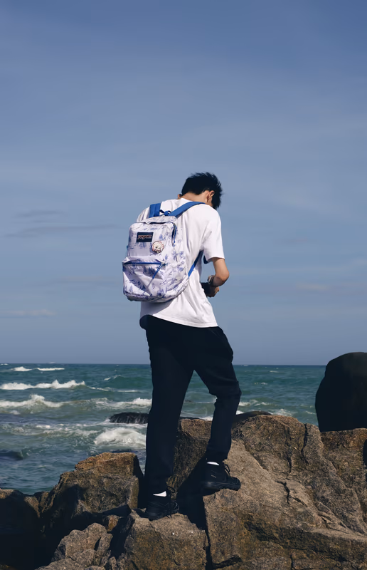 青年背着印花背包站在海边岩石上